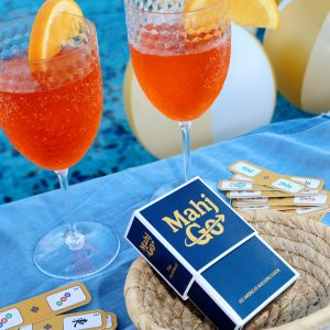 A blue MahjGo pocket-sized Mahjong playing card deck sitting next to two vibrant orange spritz cocktails with orange slices on a blue table.